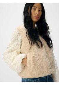 Beige fleece vest med en blød, tekstureret overflade og en afrundet kant, lagt over en gennemsigtig, flæset langærmet top med cremede nuancer.