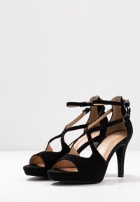 Czarne zamszowe sandały na obcasie z designem T-strap, otwartym noskiem i zapięciem na kostkę. Posiadają platformę dla dodatkowej wysokości. Gładkie wewnętrzne wykończenie.
