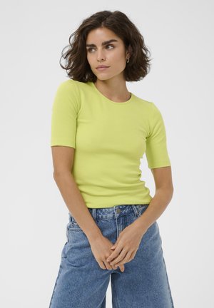 Kobieta o krótkich kręconych brązowych włosach, ubrana w limonkową koszulkę z krótkim rękawem i niebieskie, wysokie dżinsy, stojąca z splecionymi rękami.