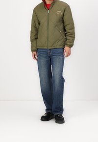 Olivengrøn quiltet jakke med høj krave, lynlås lukning og elastiske manchetter, parret med blå denim jeans og sorte sko.