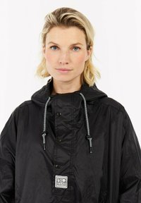 Zwarte regenjack met een trekkoord capuchon, drukknoopsluiting en een kenmerkend logopatch. Glad, lichtgewicht stof met een glanzende afwerking.