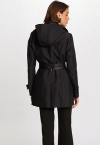 Svart trenchcoat med hette, teksturert materiale, beltestedde midje og knappeermer. Designet med en tettsittende silhuett og plisserte bak.