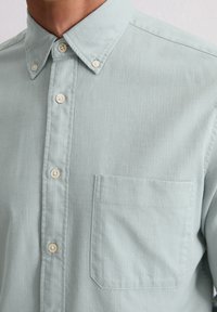 Camicia azzurro chiaro con bottoni a pressione, realizzata in tessuto testurizzato, presenta un colletto a punta, bottoni bianchi e una tasca sul petto con un'apertura quadrata.