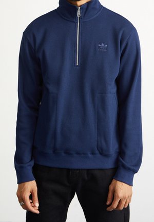 Mann trägt einen dunkelblauen langärmligen Pullover mit Halb-Reißverschluss und dezentem Adidas-Logo auf der linken Brust, kombiniert mit schwarzen Hosen.