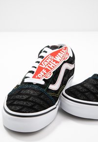 Baskets Vans noires avec lacets blancs et semelles gaufrées, avec une couture multicolore et une étiquette rouge "Vans Off The Wall" sur une chaussure.