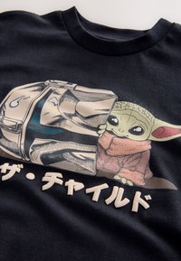 Camiseta negra de algodón con un gráfico de un personaje asomándose desde detrás de un casco metálico. Texto debajo en caracteres japoneses.