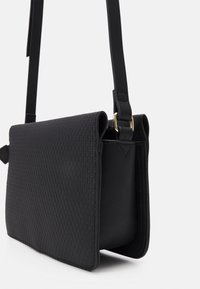 Černá kabelka přes rameno s texturovaným tkaným čelem, hladkými bocí a plochým profilem. Obsahuje zlaté kovové prvky a nastavitelné rameno.