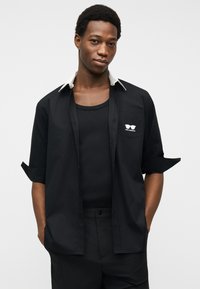 Νεαρός άνδρας που φοράει μαύρο πουκάμισο με τα μανίκια γυρισμένα, μαύρο αμάνικο και μαύρο παντελόνι, στέκεται με τα χέρια στις τσέπες μπροστά σε απλό φόντο.