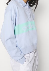 Ljusblå tröja med krage, med ett horisontellt band i mintgrönt och vit logotext. Materialet verkar mjukt och något texturerat.