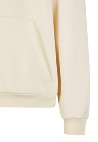 Détail de la manche de sweat-shirt beige et de la poche kangourou avec poignets et ourlet côtelés en tissu doux.