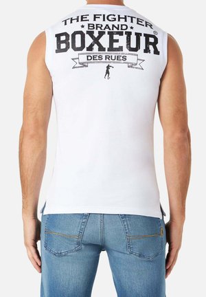 Uomo che indossa una maglietta bianca senza maniche con scritte nere "BOXEUR" e "THE FIGHTER BRAND" sul retro, abbinata a jeans blu.
