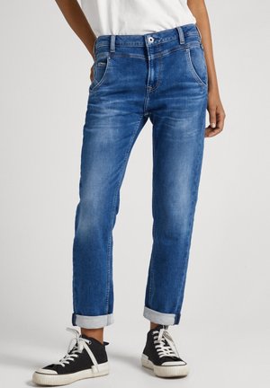 Personne portant un jean bleu retroussé, une chemise blanche et des baskets montantes noires avec des lacets blancs, se tenant devant un fond uni.