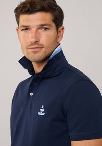 Granatowa koszulka polo z teksturowanej tkaniny, z złożonym kołnierzykiem z jasnoniebieskim wykończeniem i wyszywanym logo na piersi.