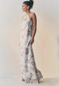 Blomstermaksikjole lavet af glat stof, med halterneck og et mønster af lyserøde roser mod en hvid baggrund. Elegant silhuet.