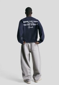 Oseba v mornarsko modrem puloverju z napisom na hrbtu in ohlapnih sivih hlačah, ki stoji obrnjen proti praznemu ozadju.