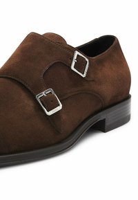 Brown suede double monk strap shoe featuring silver-tone buckles and a low black heel. Smooth texture with detailed stitching accents.
