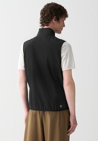 Gilet senza maniche nero con colletto alto, texture liscia e orlo elastico. Presenta un logo visibile in basso a sinistra.