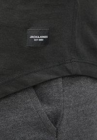 Zwarte stof met een klein zwart label met de tekst "JACK & JONES EST. 1990" bovenop een grijzige textuurstof, waarbij de textuur en naden van het kledingstuk zichtbaar zijn.
