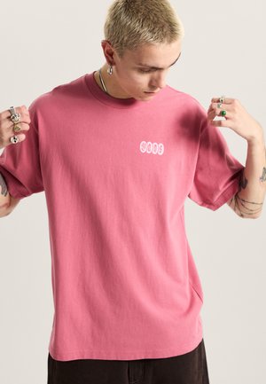 Pink cotton T-shirt with a crew neck and short sleeves. Features a white logo on the left chest area. Smooth texture, relaxed fit.