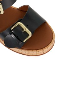 Zwarte leren sandalen met twee brede banden, gouden gespen en een kurken zool. De voetbed is van bruin leer met een gladde textuur.
