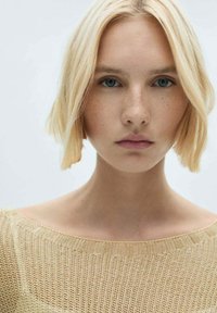 Persoană cu părul blond, având o tunsoare bob scurt, îmbrăcată într-un pulover tricotat de culoare bej deschis, cu decolteu tip barcă, prezentată pe un fundal neutru.