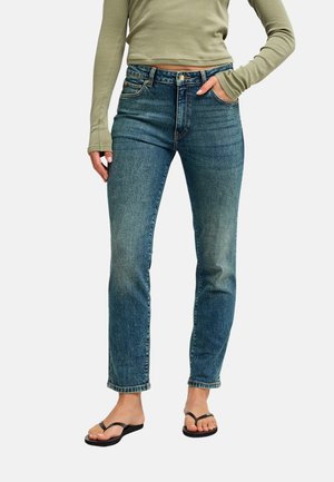 Femme portant un jean bleu taille haute, un crop top à manches longues vert olive et des sandales noires, debout avec une main dans la poche.