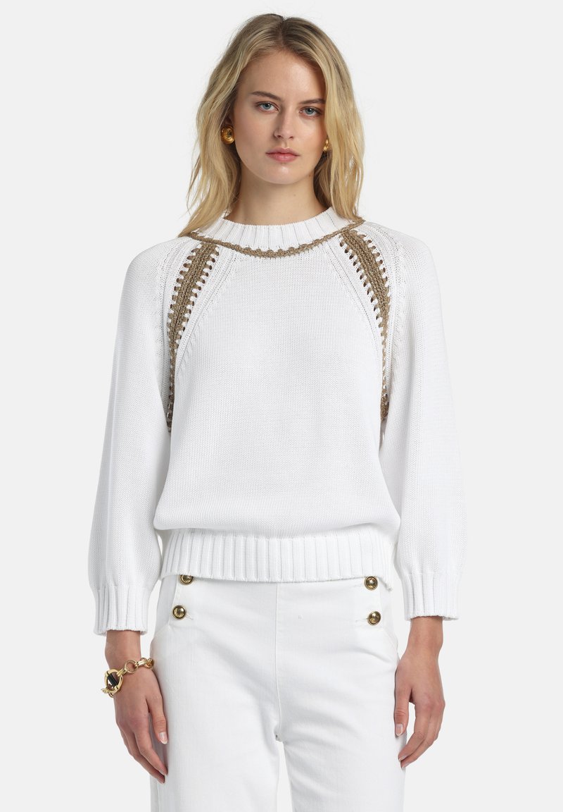 Pulover alb cu tivuri și manșete zimțate, decorat cu accente împletite aurii pe umeri și croială lejeră, purtat cu pantaloni albi.