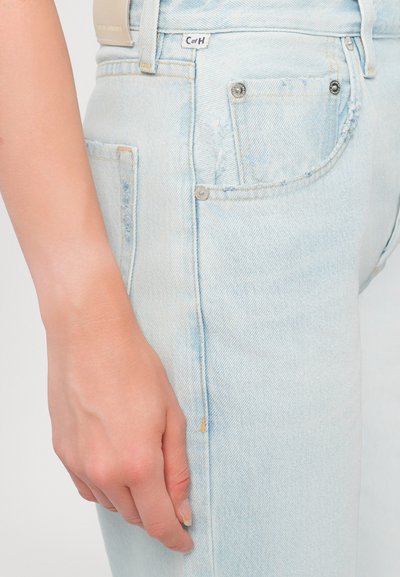 I jeans in denim blu chiaro presentano un leggero sfilacciamento, cuciture visibili e una piccola etichetta applicata in vita con due tasche anteriori.