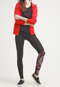 Czerwona bluza z kapturem na zamek błyskawiczny w połączeniu z czarnymi legginsami z wzorem na lewej nodze. Czarne buty wsuwane dopełniają stylizację. Prosty i casualowy design.