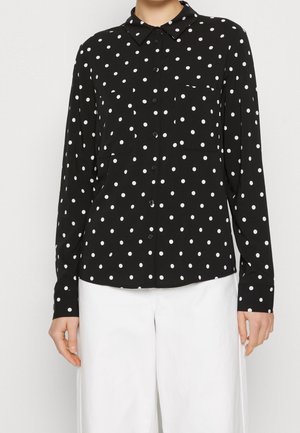 Chemise noire à manches longues boutonnée avec des pois blancs, associée à un pantalon blanc à jambes larges portée par une personne debout.
