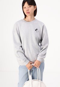 Szürke pulóver hosszú ujjakkal és kerek nyakkal, melyen egy kis fekete-fehér Mickey Mouse logó található a mellrészen. Világoskék farmernadrággal viselve.