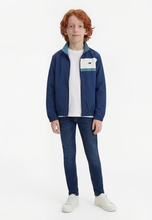 Rødhåret barn stående iført en blå lynlåsjakke over en hvid skjorte, mørke jeans og hvide sneakers mod en ensfarvet hvid baggrund.