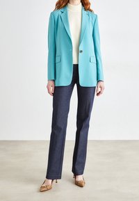 Tyrkysový blazer s přiléhavým střihem a klopovými límci, v kombinaci s tmavě modrými kalhotami a svetrem světlé barvy. Jednoduché barvy, hladká textura.