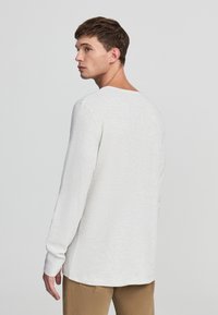 Maglietta grigia chiara a maniche lunghe con tessuto testurizzato e scollo rotondo; esposta da dietro, abbinata a pantaloni beige.