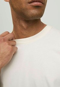 Krämbefärgad bomull t-shirt med rund hals, med en mjuk yta. Närbild av halsringningen och en del av personens hand.