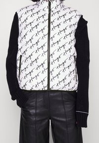 Bílá péřová vesta s černým vzorem loga ve formě písma, zeleným zipem a černými pletenými rukávy, doplněná o černé kožené kalhoty.