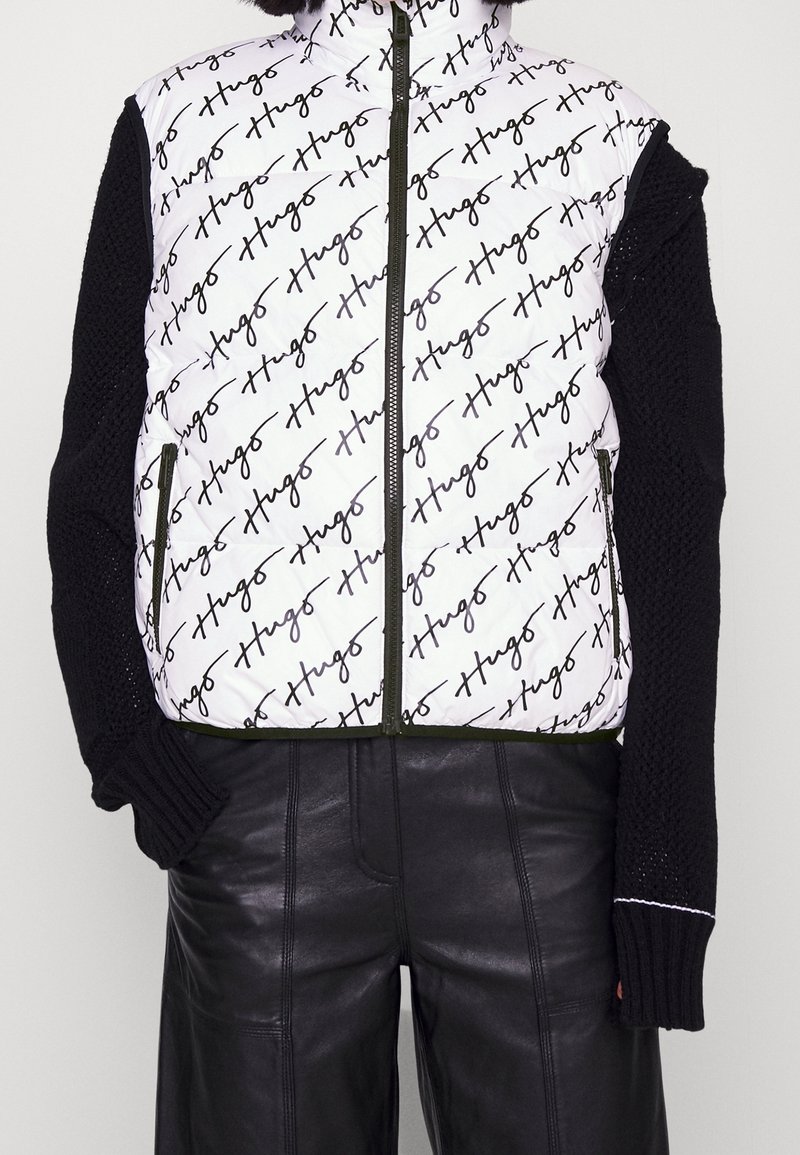 Bílá péřová vesta s černým vzorem loga ve formě písma, zeleným zipem a černými pletenými rukávy, doplněná o černé kožené kalhoty.