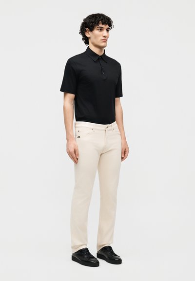 Άνδρας με σγουρά μαλλιά στέκεται φορώντας μαύρο πόλο με κοντά μανίκια, κρεμ παντελόνι στενής γραμμής και μαύρα αθλητικά παπούτσια μπροστά σε απλό ανοιχτό φόντο.