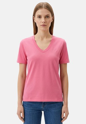 Donna che indossa una maglietta rosa semplice a maniche corte con scollo a V e jeans blu, in piedi davanti a uno sfondo grigio chiaro.