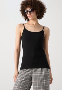 Schwarzes, figurbetontes Trägershirt mit dünnen Trägern, kombiniert mit grauen und rosa karierter Hose, vor einem schlichten Hintergrund getragen. Textilstruktur.