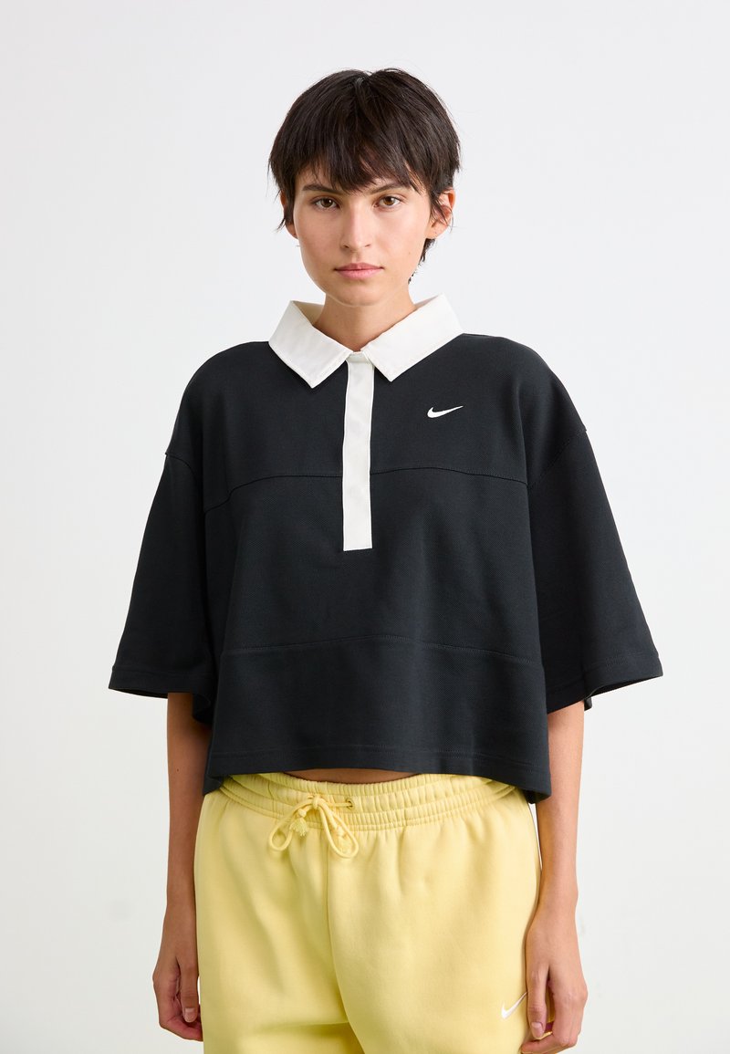 Čierny skrátený pólový tričko s krátkymi rukávmi, bielym golierom a logom Nike na hrudi. V kombinácii so žltými teplákmi so sťahovacou šnúrkou.