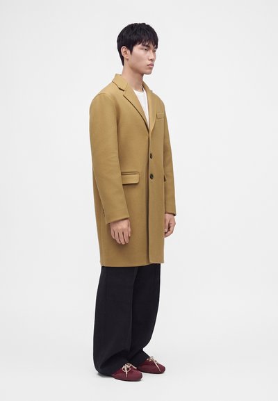 Homme debout de profil portant un long manteau beige, un pantalon large noir, une chemise blanche et des chaussures rouges avec des lacets beiges sur fond blanc uni.