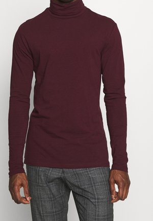 Mann trägt einen eng anliegenden burgunderroten Rollkragenpullover und graue karierte Hose, steht vor einem schlichten weißen Hintergrund.