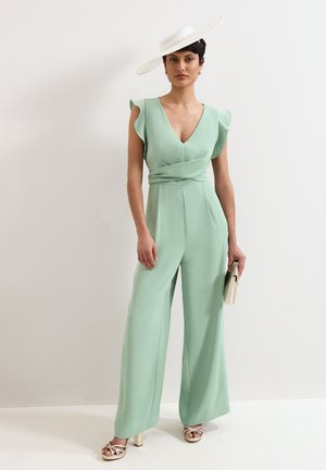 Vrouw die een lichtgroene mouwloze jumpsuit draagt met ruches op de schouders, een witte hoed met brede rand, een beige clutch en sandalen met hoge hakken.