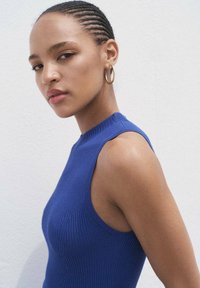 Blue ribbed sleeveless top with a high neckline. Model wears a gold hoop earring, featuring a smooth texture against a white background.