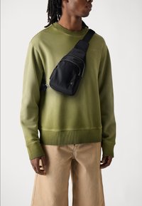 Černá taška přes rameno s několika zipovými kapsami, nošená přes olivově zelenou mikinu. Světle zbarvené široké kalhoty dokončují outfit.