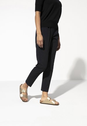Sandalias doradas tipo sandalia con dos correas ajustables, plantilla de corcho y suela de goma. Combinadas con pantalones oscuros y entallados y una blusa negra.