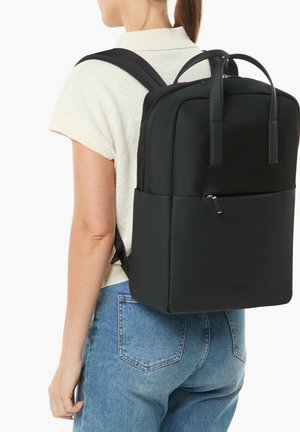 Woman wearing a black rectangular backpack with top handles, zippered front pocket, and adjustable shoulder straps, dressed in light top and blue jeans.