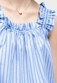 Κοντινό πλάνο ενός ατόμου που φοράει μπλε και λευκή ριγωτή μπλούζα χωρίς μανίκια με βολάν και χρυσό μενταγιόν.