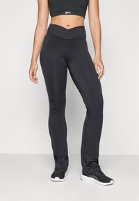 Black fitted workout leggings made of stretch material, featuring a high, crossover waistband and smooth texture. Paired with black sports shoes.